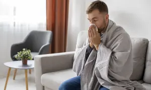 Mężczyzna siedzi na kanapie, otulony kocem, z chusteczką przy nosie. Czy to objawy przeziębienia, czy może zespół Munchausena?