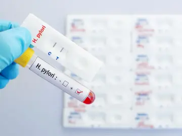 Ręka w rękawiczce trzyma probówkę z oznaczeniem "H. pylori" i zaznaczonym wynikiem dodatnim, obok test kasetkowy z dwoma paskami.