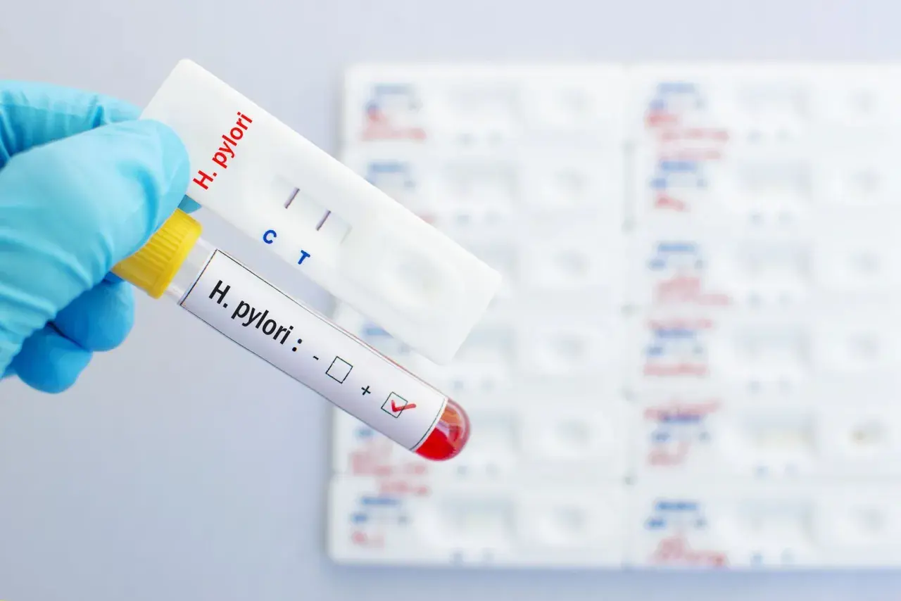 Ręka w rękawiczce trzyma probówkę z oznaczeniem "H. pylori" i zaznaczonym wynikiem dodatnim, obok test kasetkowy z dwoma paskami.
